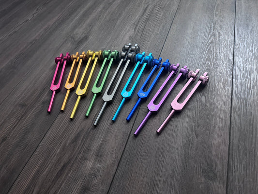 May include: A set of 12 tuning forks in various colors, including pink, yellow, green, blue, purple, and silver. The tuning forks are arranged in a row on a dark wooden surface.
