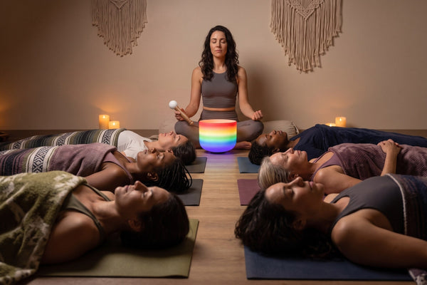 A practitioner seated at the head of a dim candlelit studio holding a mallet beside a glowing rainbow crystal singing bowl, with participants lying under blankets in savasana during a group sound bath