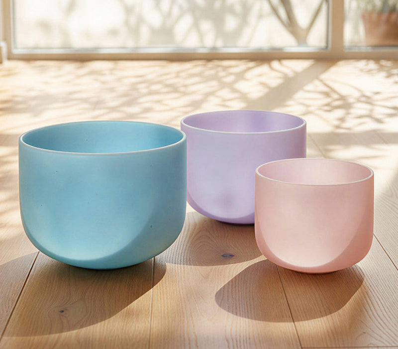 Three pastel-colored crystal singing bowls arranged on a wooden surface in natural sunlight, creating a calm and peaceful sound healing atmosphere.