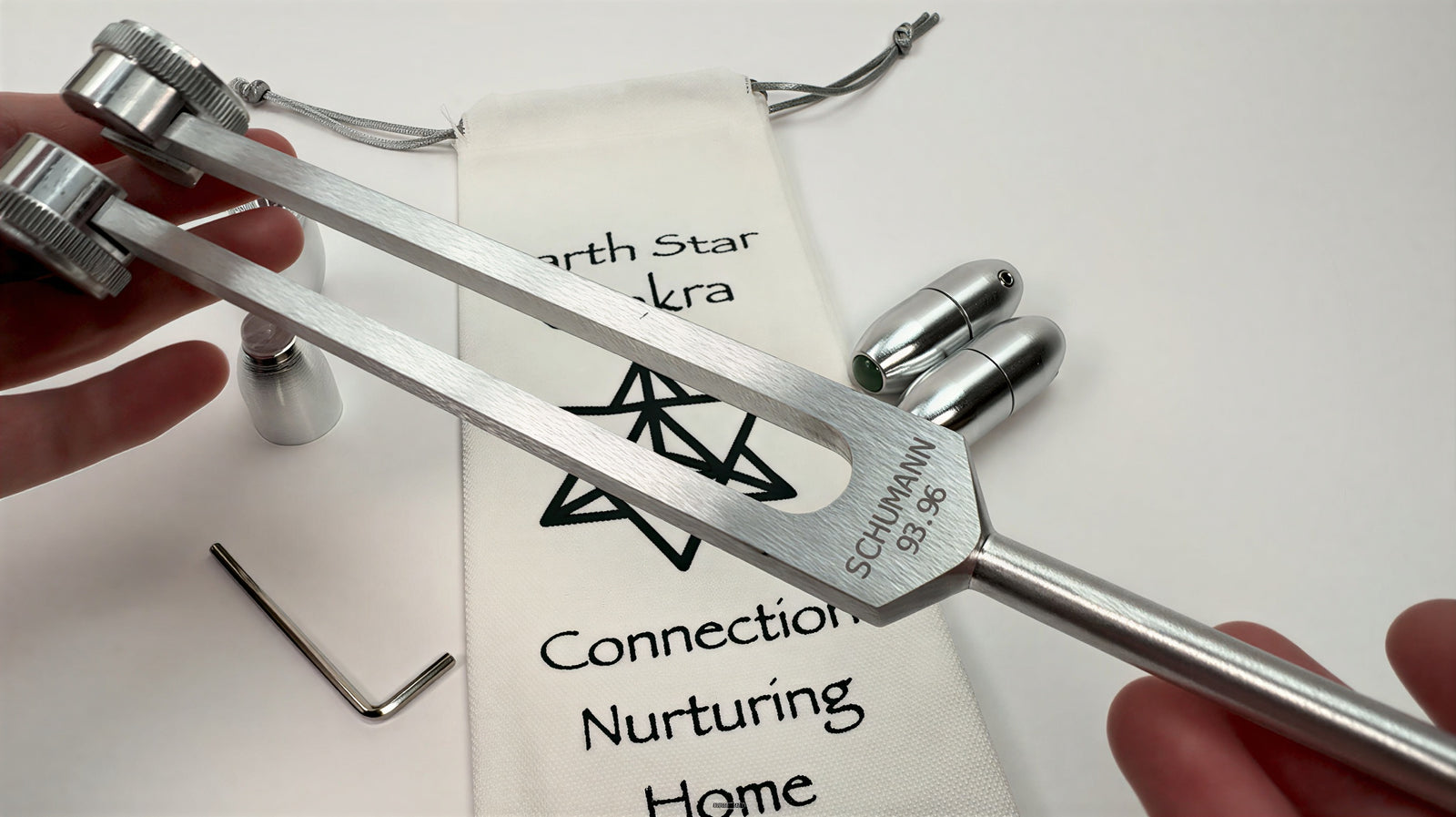 Close-up demonstration of a Schumann 7.83 Hz tuning fork with metal attenuator attachments, held above a branded pouch during a sound healing and frequency meditation practice.