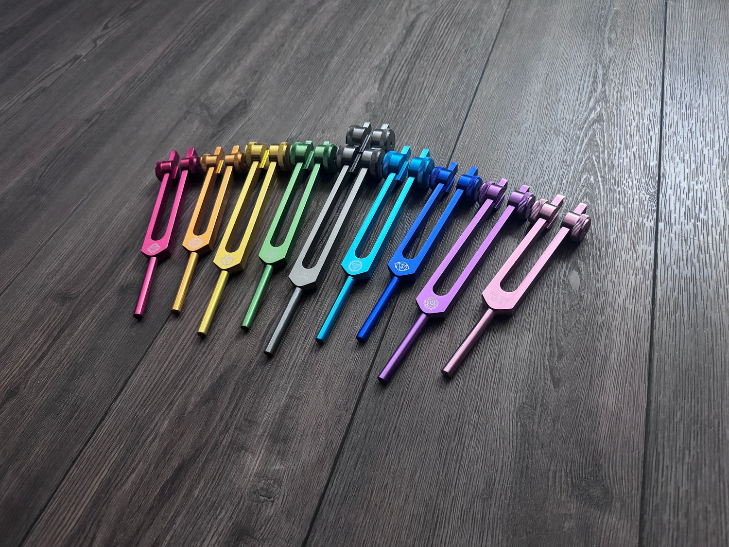 May include: A set of 12 tuning forks in various colors, including pink, yellow, green, blue, purple, and silver. The tuning forks are arranged in a row on a dark wooden surface.