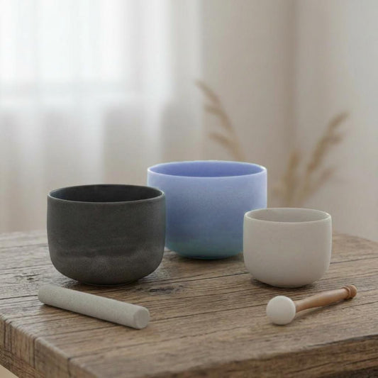 May include: Three sound bowls in shades of gray, blue, and white sit on a wooden surface. A gray mallet and a wooden mallet with a white head are also present. The bowls are likely used for sound healing or meditation.